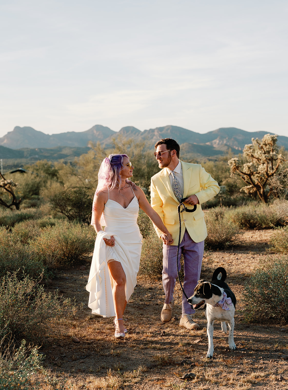 Bride and groom walking together in the desert with their dog, smiling at each other during golden hour