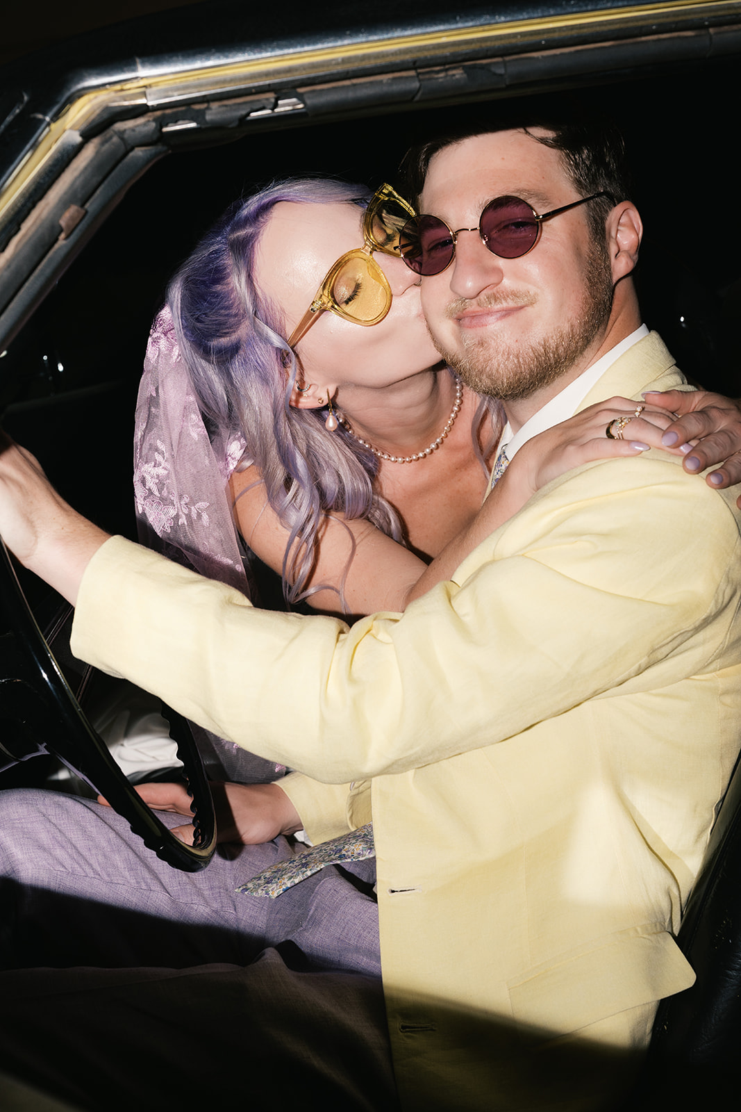 Bride kissing groom while sitting together in a vintage car, both wearing sunglasses and smiling