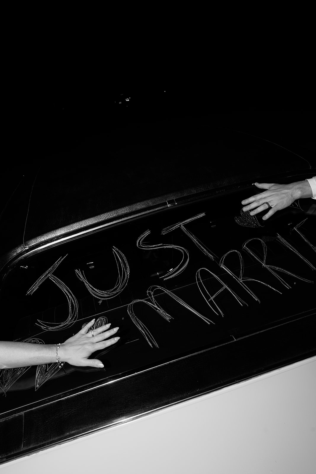 Hands writing “just married” on the back of a car window in a playful black and white moment