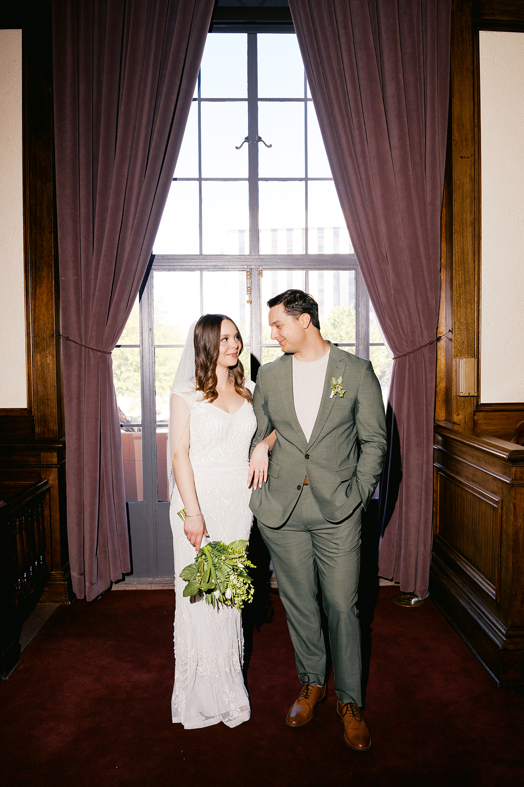 Non-traditional wedding couple posing by tall curtains indoors