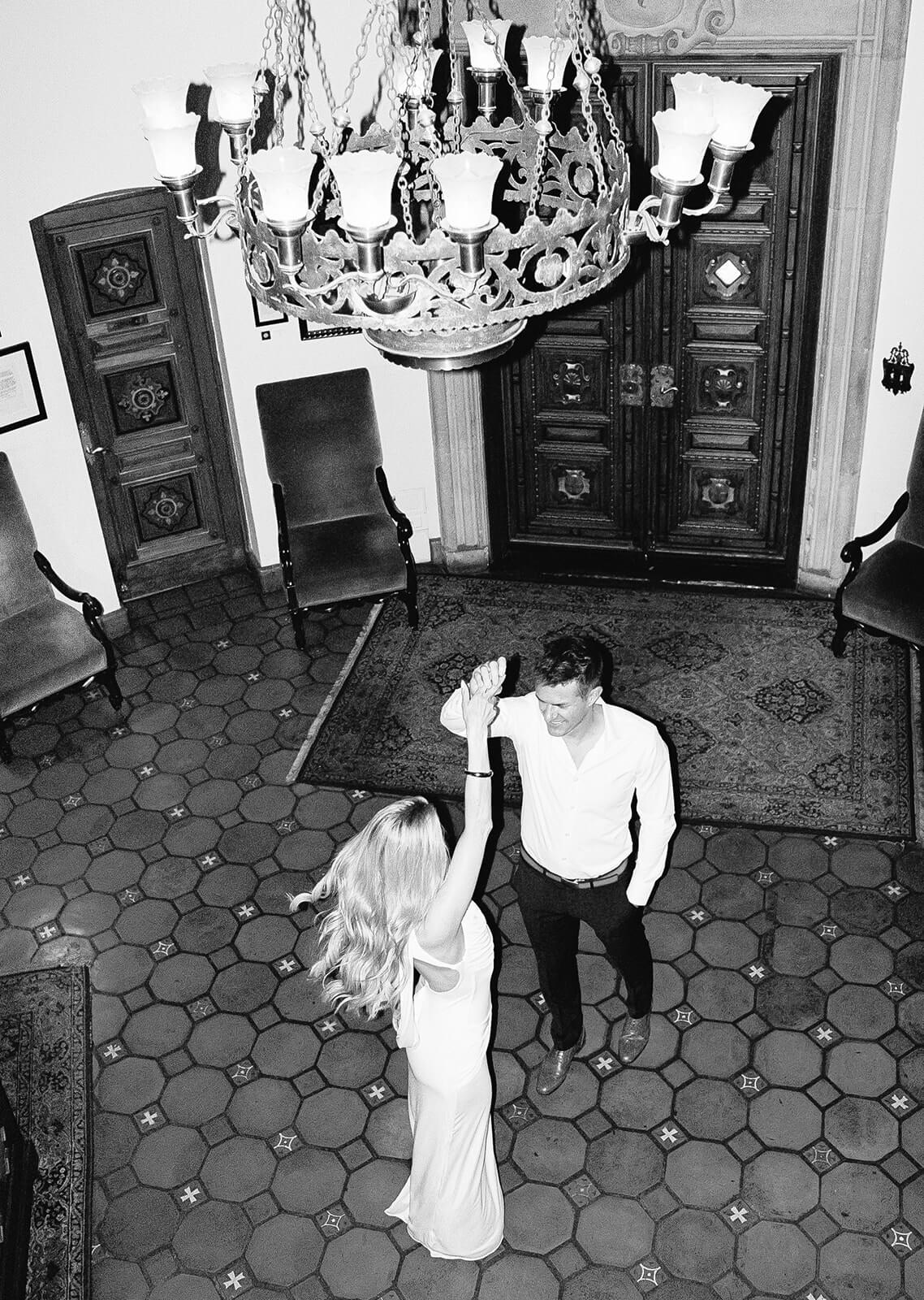 Black-and-white overhead photo of a couple dancing beneath an ornate chandelier in a vintage-style room. The woman in a long dress raises her arm as if twirling, while the man in a button-down shirt and dark pants looks up at her. The space features patterned tile flooring, antique wooden doors, and classic furniture, creating a timeless, elegant atmosphere.