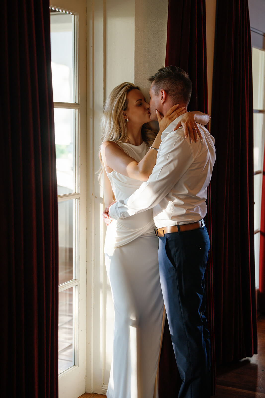 Bride and groom sharing a kiss by a sunlit window with deep red curtains at the Wrigley Mansion in Phoenix, Arizona, creating an intimate and romantic wedding moment.