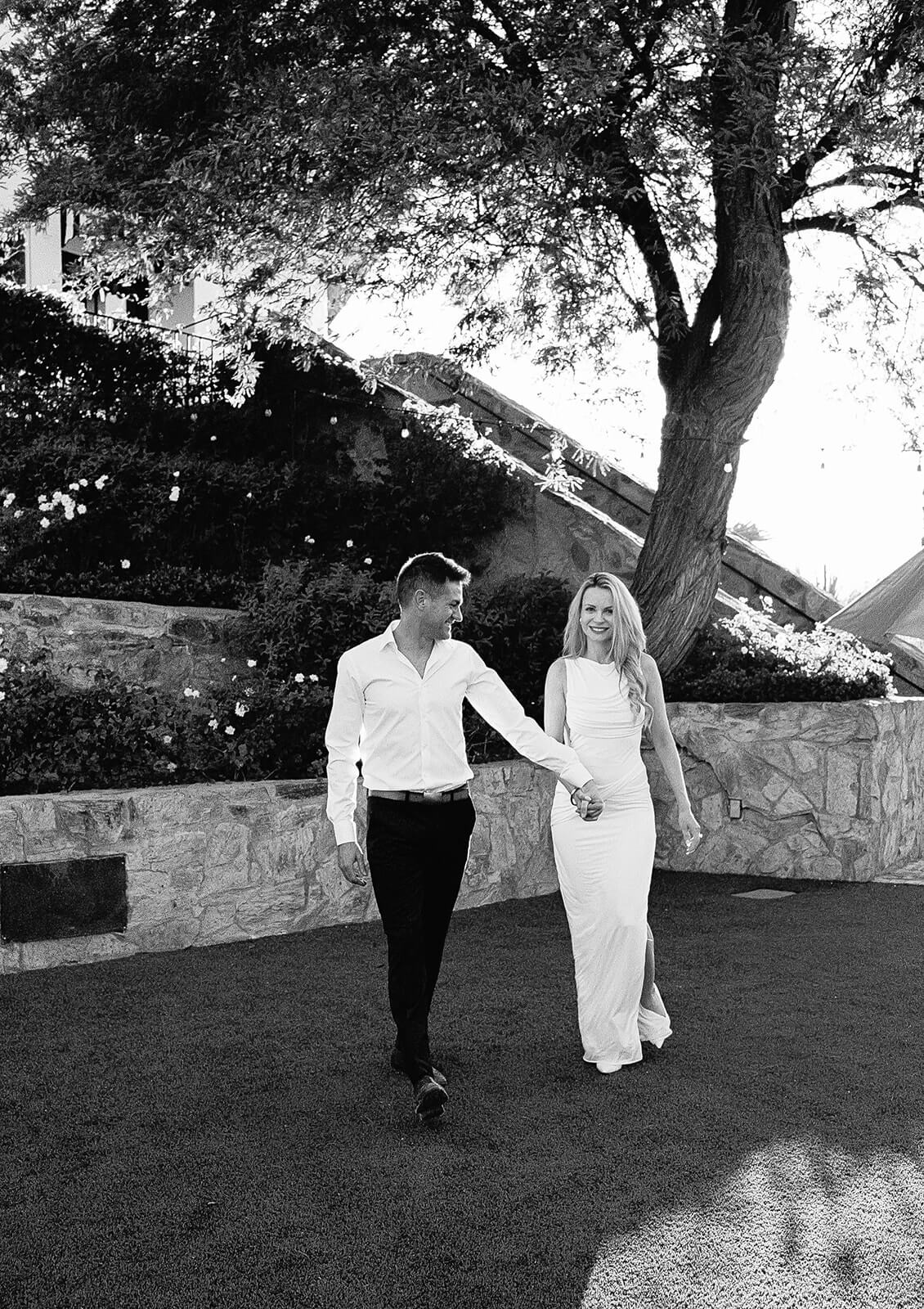 Black and white image of a couple walking hand in hand outdoors at the Wrigley Mansion, a popular Arizona wedding venue, surrounded by trees and soft natural light.