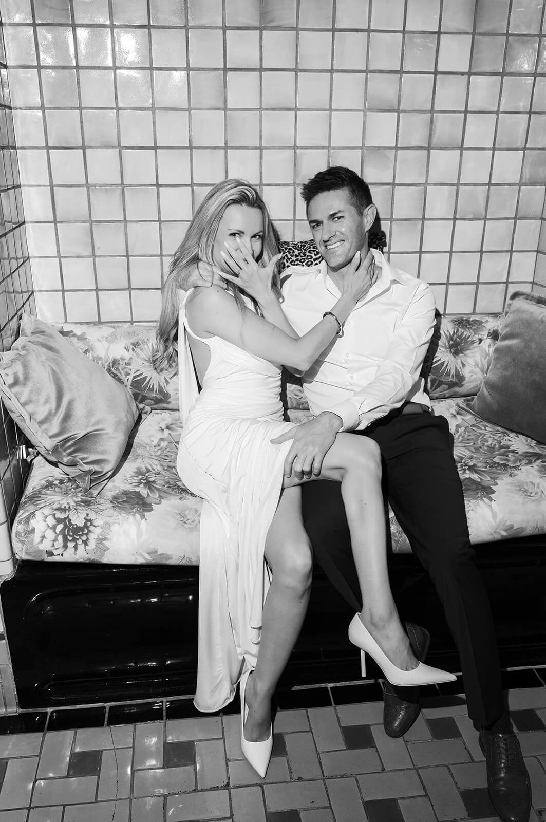 Playful black and white photo of a bride showing off her ring while sitting with her partner on a patterned couch at the Wrigley Mansion, a unique Arizona wedding venue.