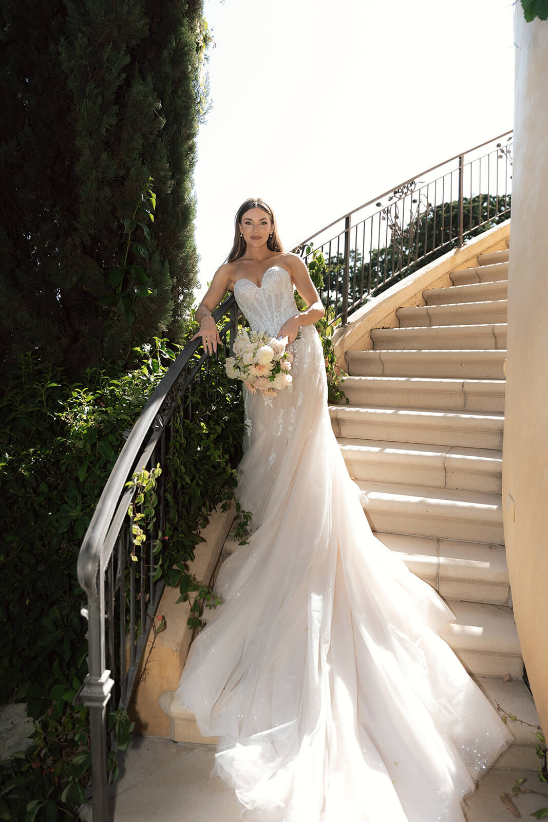 Bride posing on a staircase at a San Luis Obispo wedding venue, holding a bouquet with sunlight highlighting her detailed lace gown.
