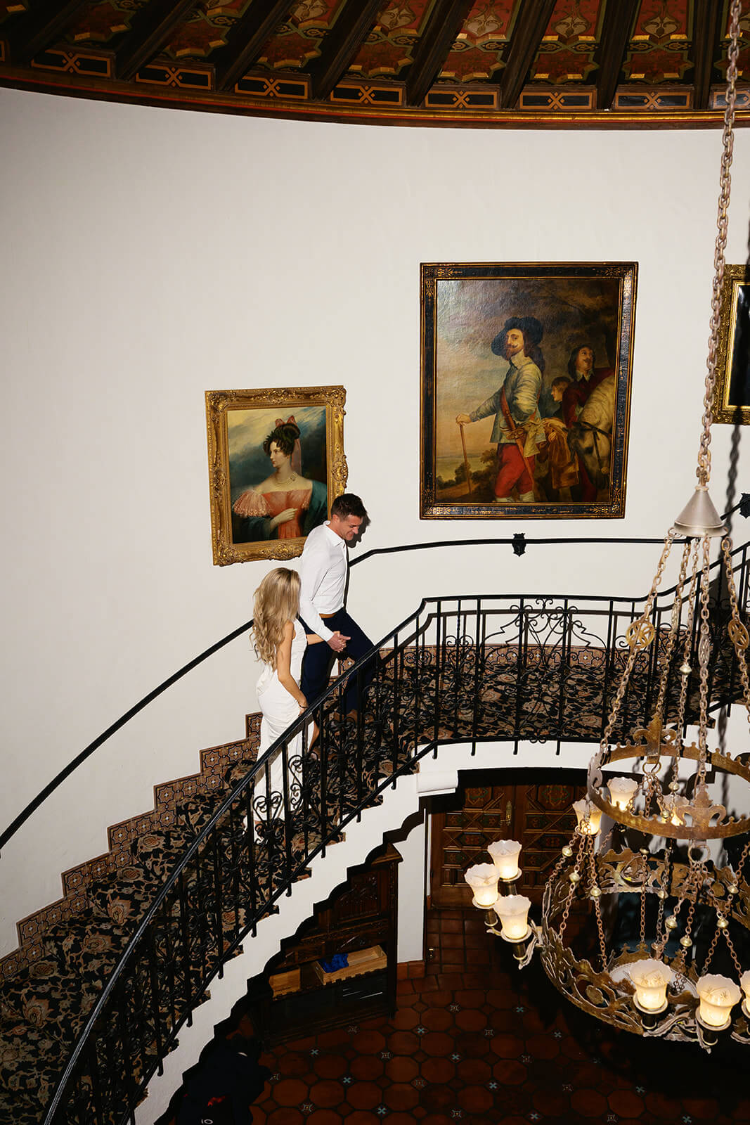 Couple walking down a grand curved staircase inside Wrigley Mansion wedding venue in Phoenix Arizona with ornate iron railing and vintage paintings