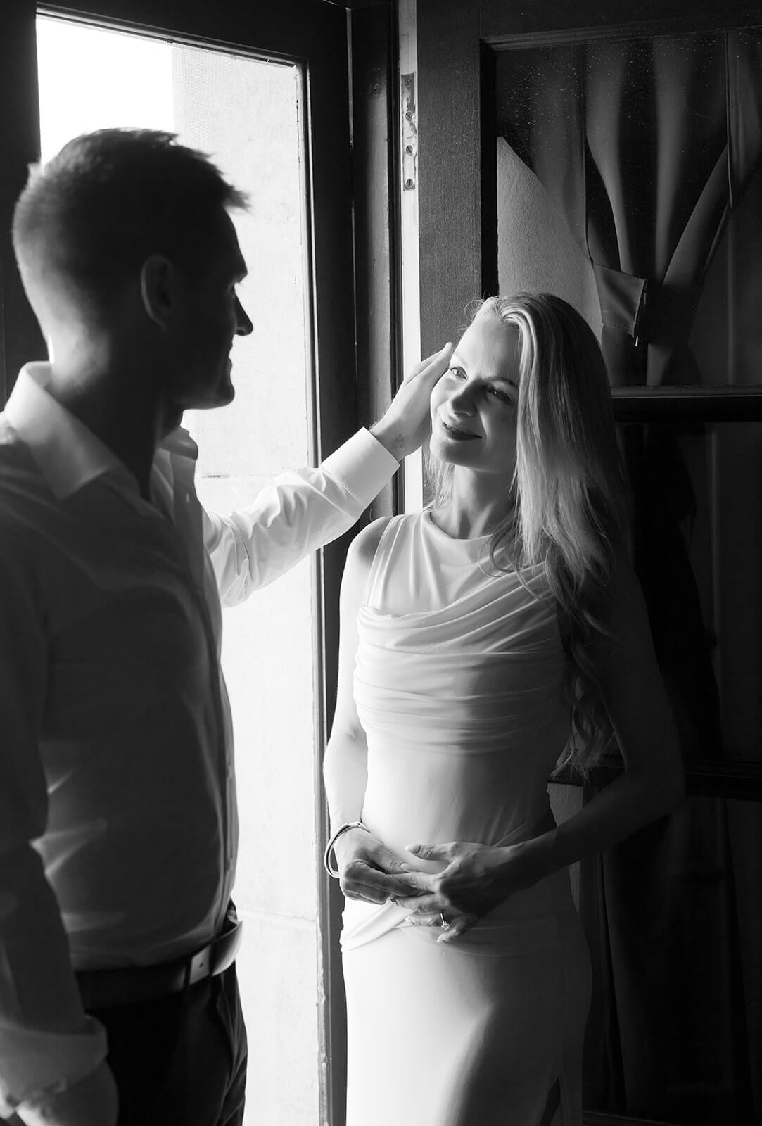 Black-and-white photo of a couple standing by a doorway, softly backlit by natural light. The man gently touches the woman’s hair as she smiles up at him, wearing a fitted dress. Their relaxed posture and the contrast between light and shadow create an intimate, quiet moment.