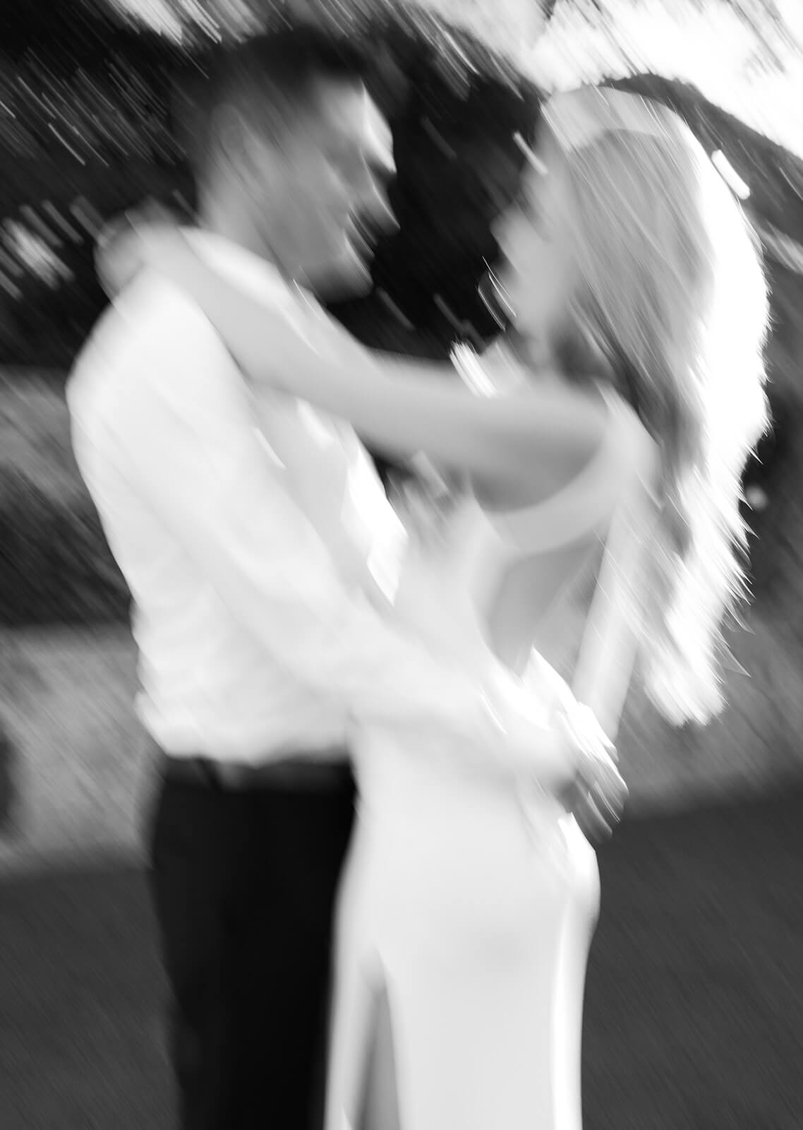 Soft, motion-blurred black-and-white photo of a couple embracing outdoors, their faces close as if about to kiss. The woman has her arms around the man’s neck, and both figures are intentionally out of focus, creating a dreamy, romantic feel with light filtering through trees in the background.