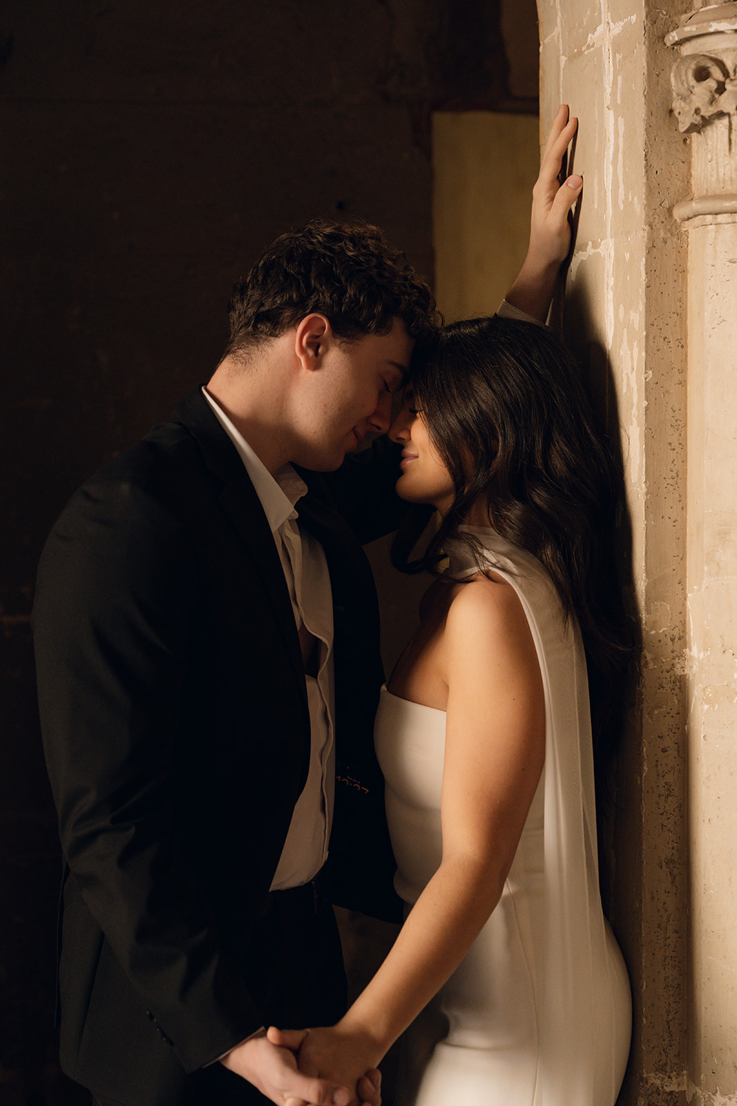 Groom leaning against a stone archway with his arm above the bride as they smile at each other in warm indoor lighting at wedding venues in Phoenix Arizona— The Abbey on Monroe