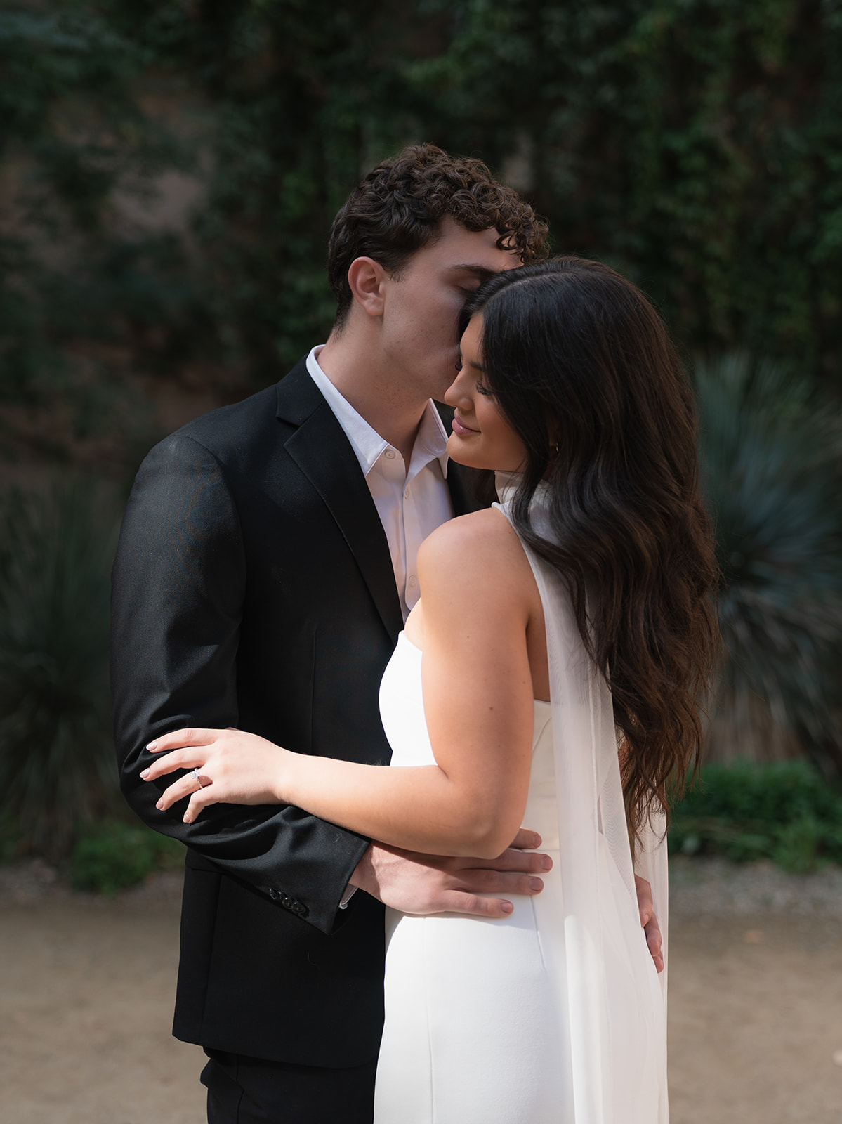 Groom kisses a bride on the temple as she smiles and wraps her arms around him in a garden courtyard at wedding venues in Phoenix Arizona— The Abbey on Monroe