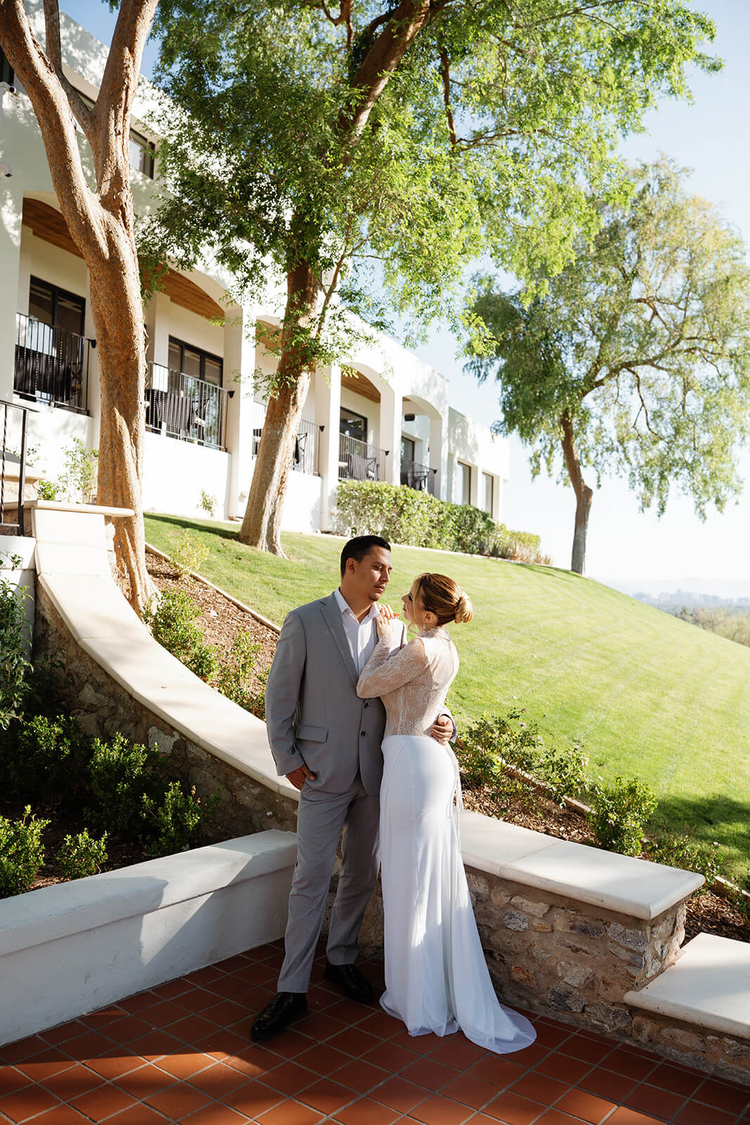 Bride and groom standing close together on a curved stone walkway at a romantic micro wedding venue, surrounded by trees and Spanish-style architecture.