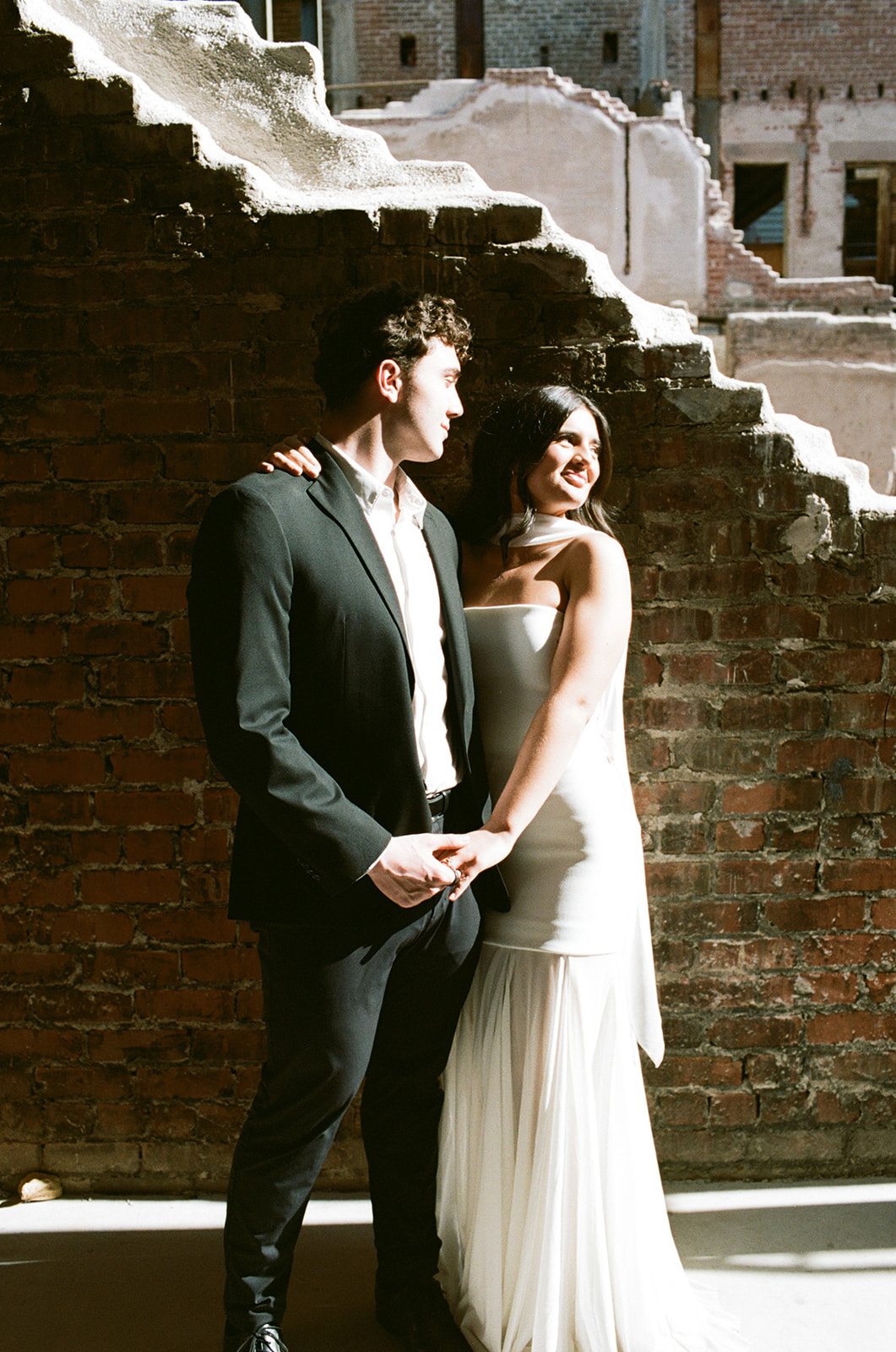 Bride leaning into the groom with her arm around his shoulder as they hold hands and smile in warm sunlight against a textured brick wall at outdoor wedding venues in phoenix — The Abbey on Monroe