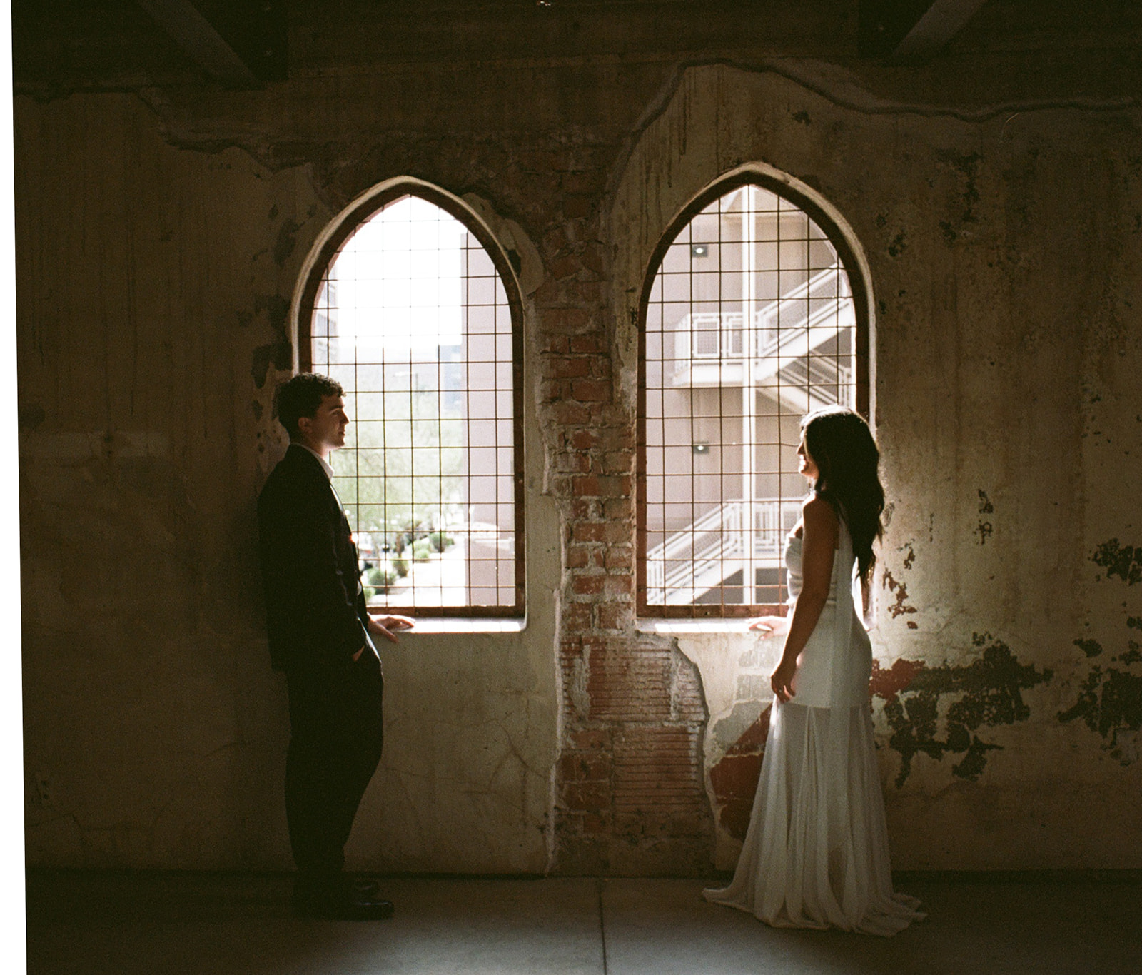 Bride and groom standing across from each other between two arched windows with metal grates, softly backlit by natural light against textured, weathered walls at outdoor wedding venues in phoenix — The Abbey on Monroe