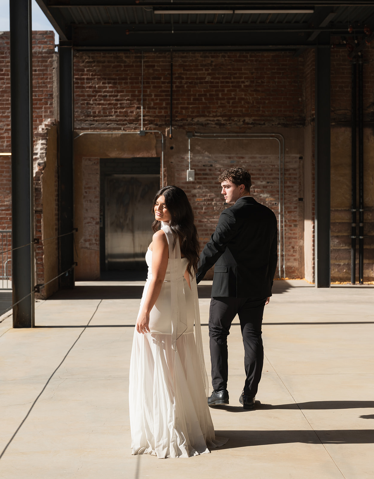 Bride and groom holding hands as they walk forward across an industrial courtyard, both looking back over their shoulders in bright sunlight at outdoor wedding venues in phoenix — The Abbey on Monroe