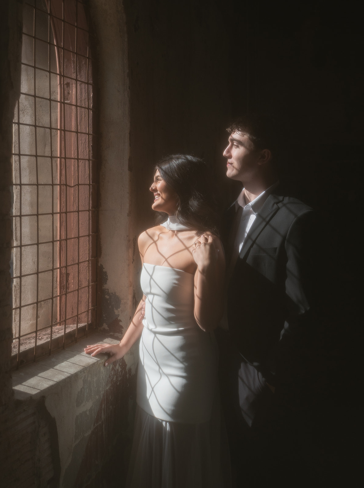 Bride smiling as she looks out a window while the groom stands just behind her with his arm around her waist, both softly lit by warm natural light and window shadows at outdoor wedding venues in phoenix — The Abbey on Monroe