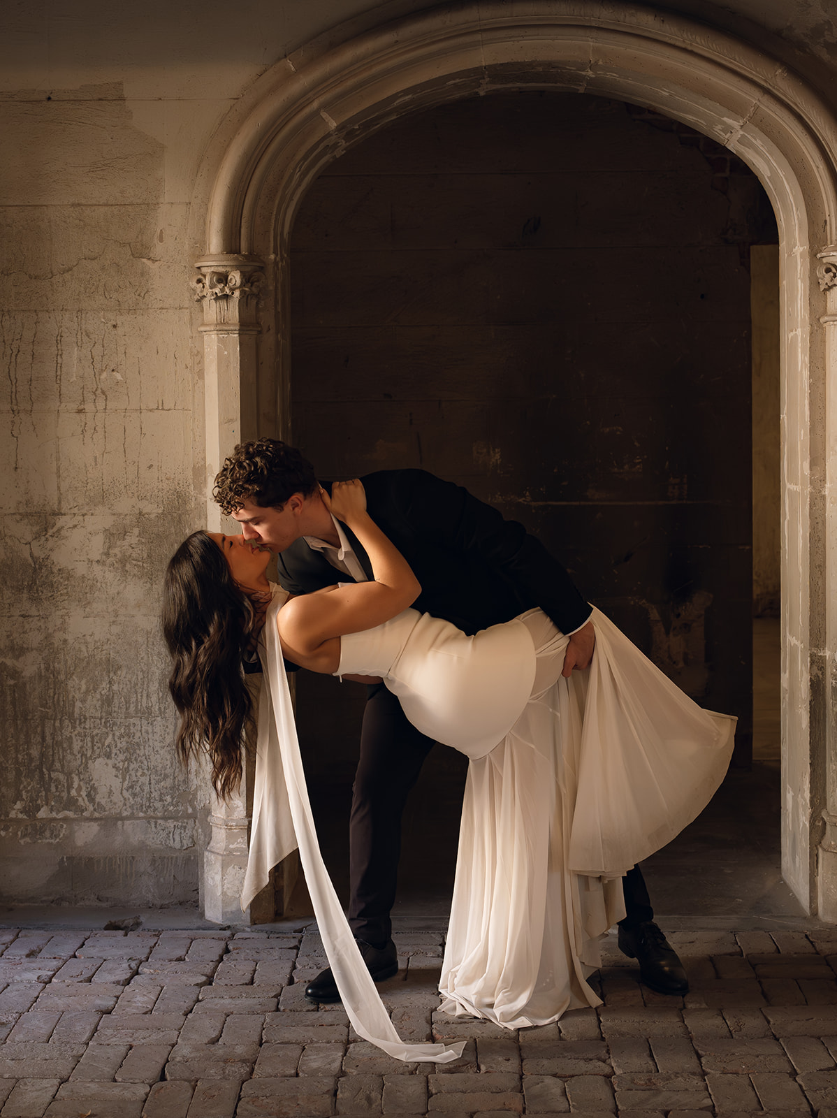 Groom dipping the bride as they kiss beneath a stone archway with warm moody lighting and textured walls at outdoor wedding venues in phoenix — The Abbey on Monroe