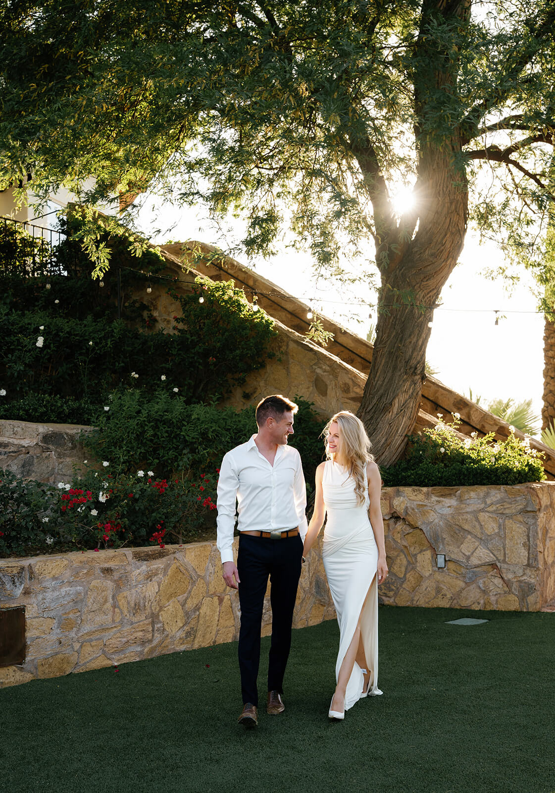 Bride and groom walking toward the camera on a narrow walkway outside a romantic micro wedding venue, surrounded by lush landscaping.