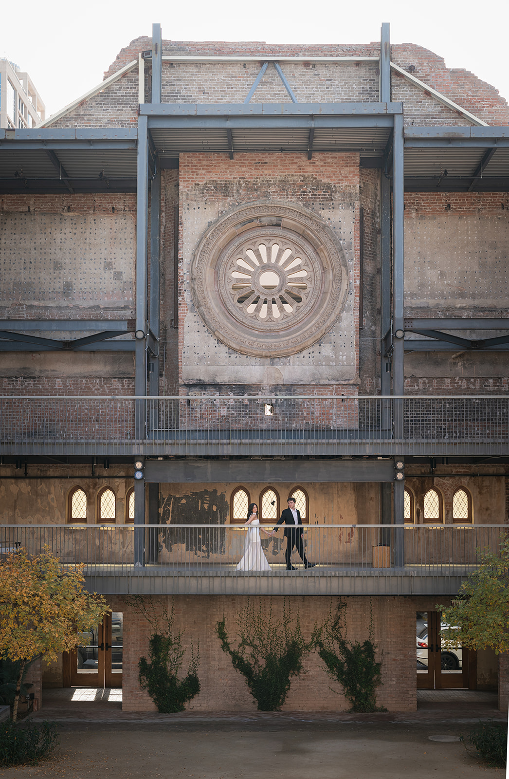 Wide shot of bride and groom holding hands while walking across an upper balcony of a historic brick building with a large circular window at outdoor wedding venues in phoenix — The Abbey on Monroe