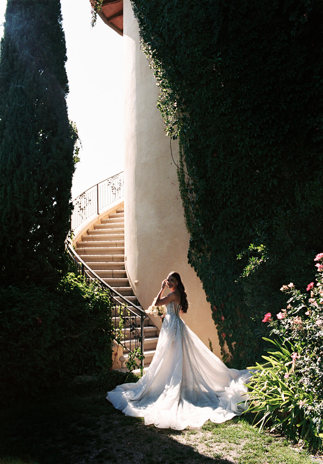 Bride standing in a sunlit garden walkway at a San Luis Obispo wedding venue, with a long flowing train and curved staircase framed by lush greenery.