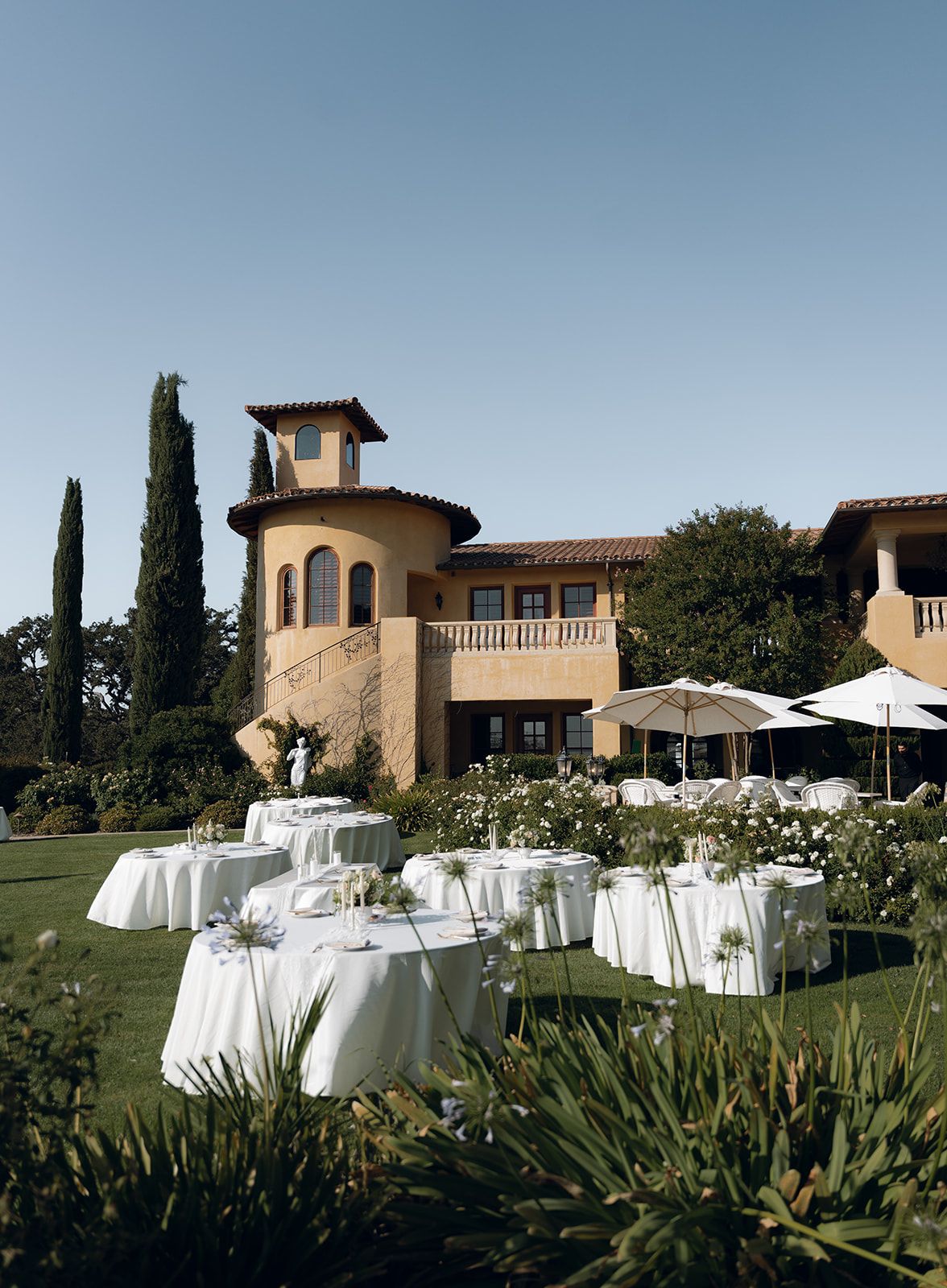 Elegant outdoor wedding reception setup at Villa San Juliette, featuring round tables with white linens arranged on a manicured lawn, surrounded by blooming greenery and overlooking a Tuscan-style villa in San Luis Obispo
