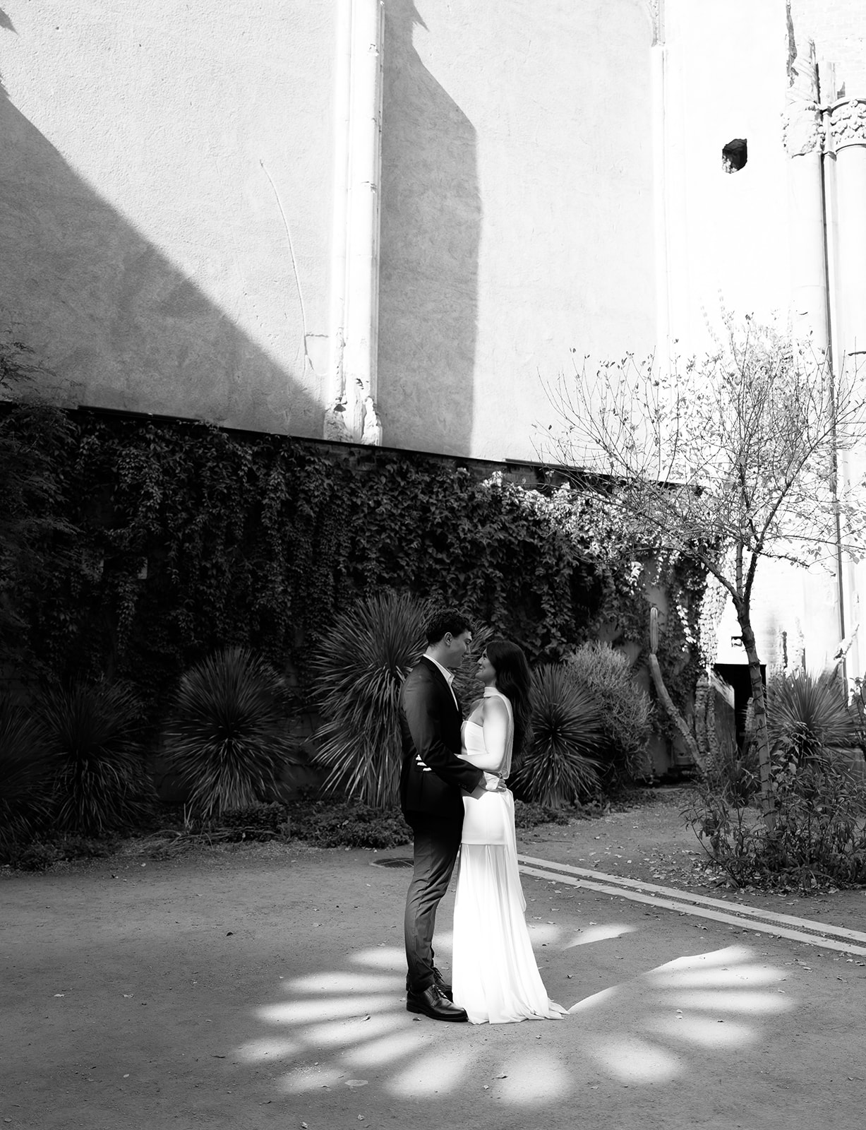 Black and white photo of bride and groom standing close together and embracing in a courtyard with desert plants and dramatic sunlight patterns on the ground at small wedding venues in phoenix az— The Abbey on Monroe