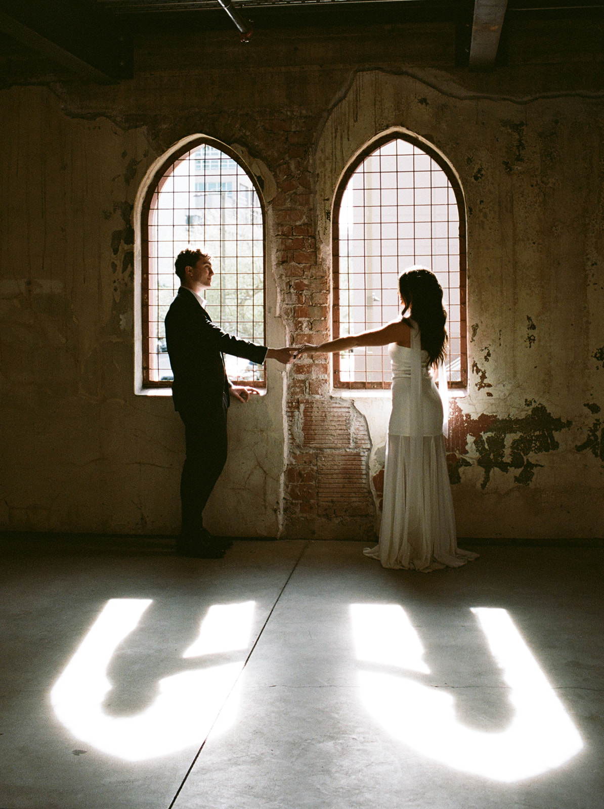 Bride and groom standing apart and holding hands in front of tall arched windows with sunlight casting shadows on the floor at small wedding venues in phoenix az— The Abbey on Monroe