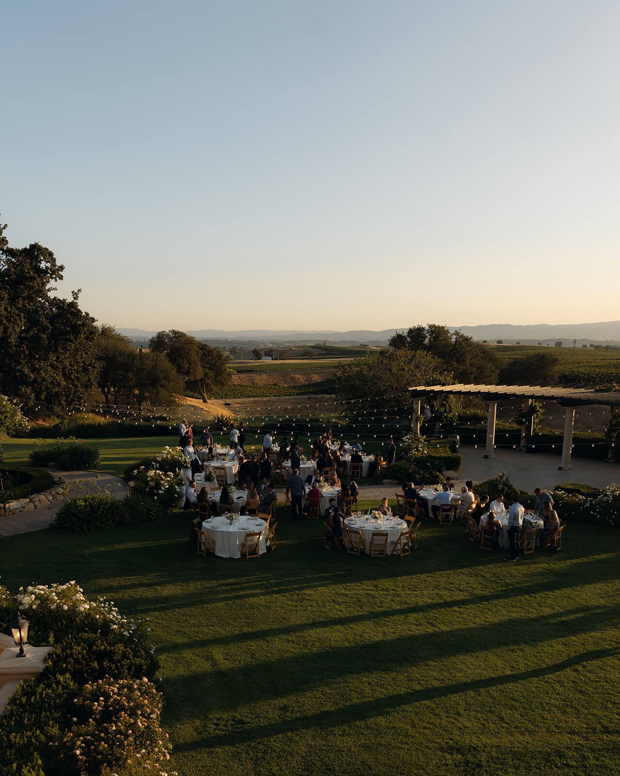 Outdoor wedding reception at a San Luis Obispo wedding venue, with guests seated at round tables on a lawn overlooking rolling hills at sunset.