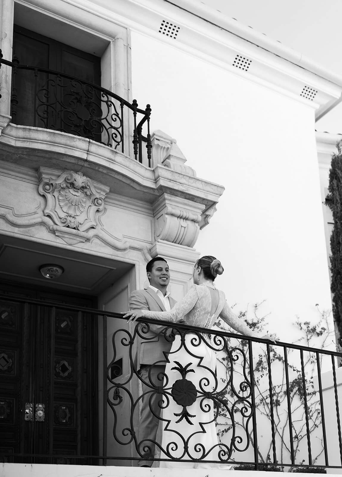 Black and white photo of bride and groom standing on a balcony at a romantic micro wedding venue, facing each other against ornate architectural details.