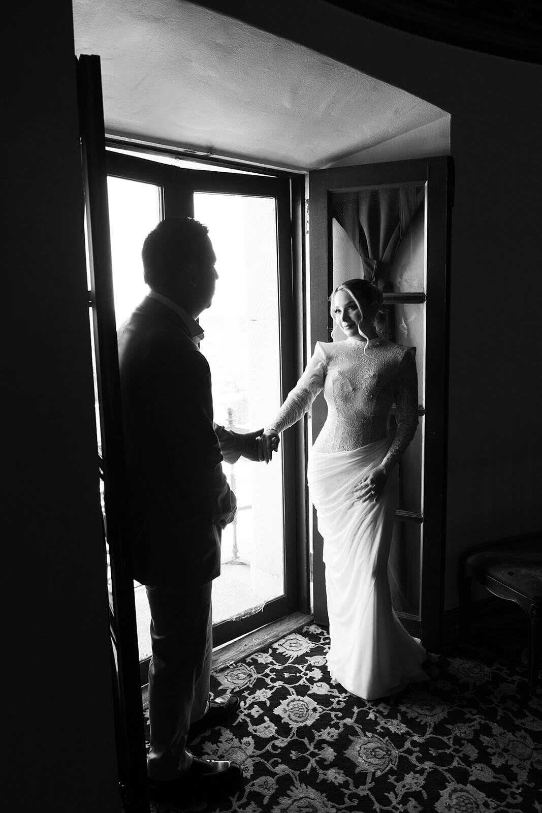 Black and white image of couple holding hands near a doorway, backlit by natural light inside an intimate micro wedding venue.