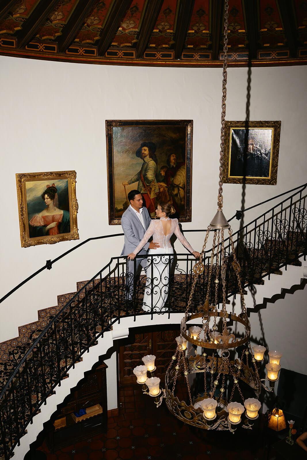 Color image of couple embracing on a curved staircase inside a historic micro wedding venue, with dramatic lighting and ornate interior details.