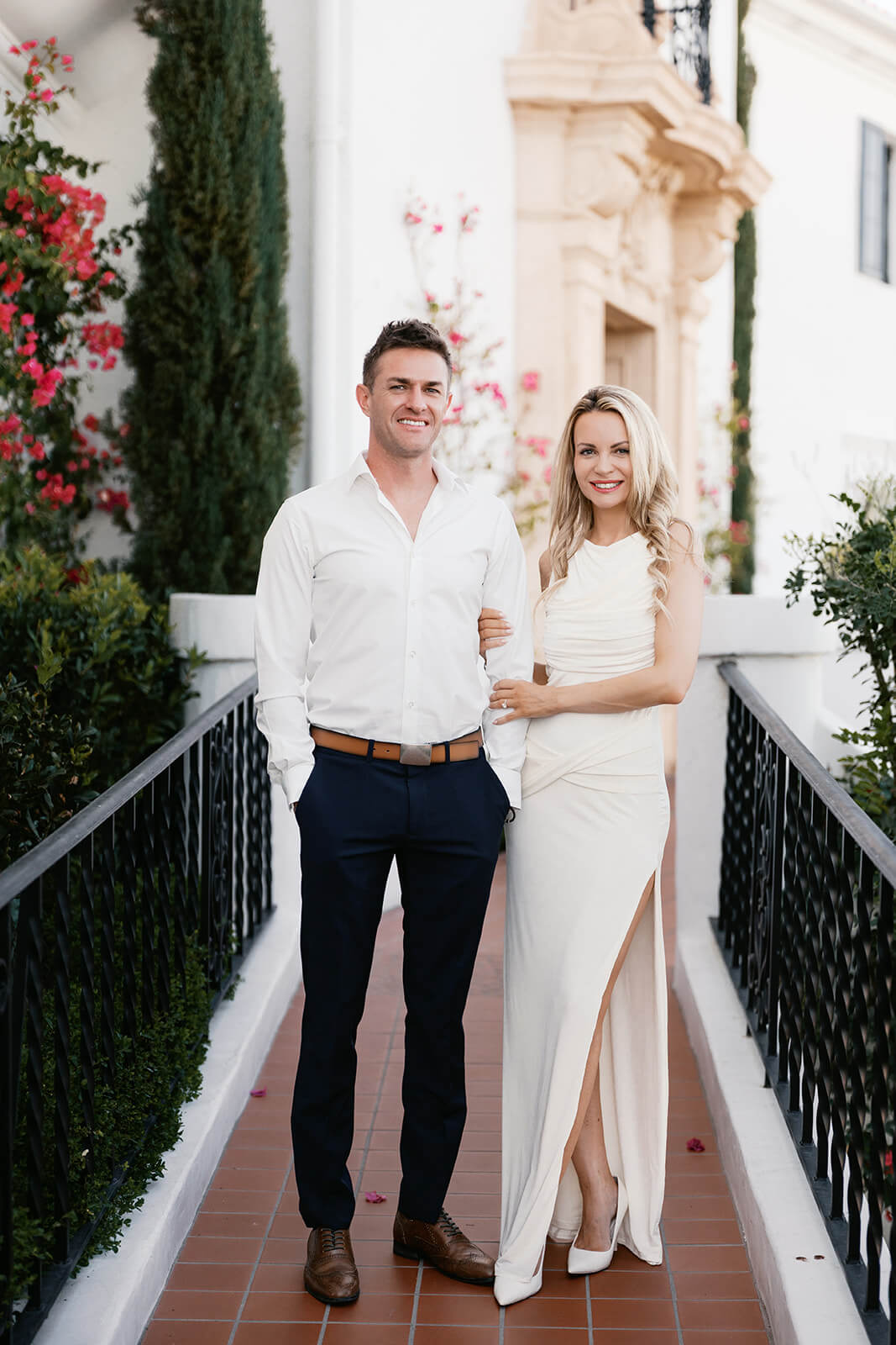 Couple walking arm-in-arm along a narrow tiled pathway, dressed in coordinated white outfits, with the woman wearing a fitted dress with a high slit and heels and the man in a white button-down shirt and dark pants. They are smiling at the camera, framed by black railings, lush greenery, and pink flowering vines against a light-colored building backdrop.