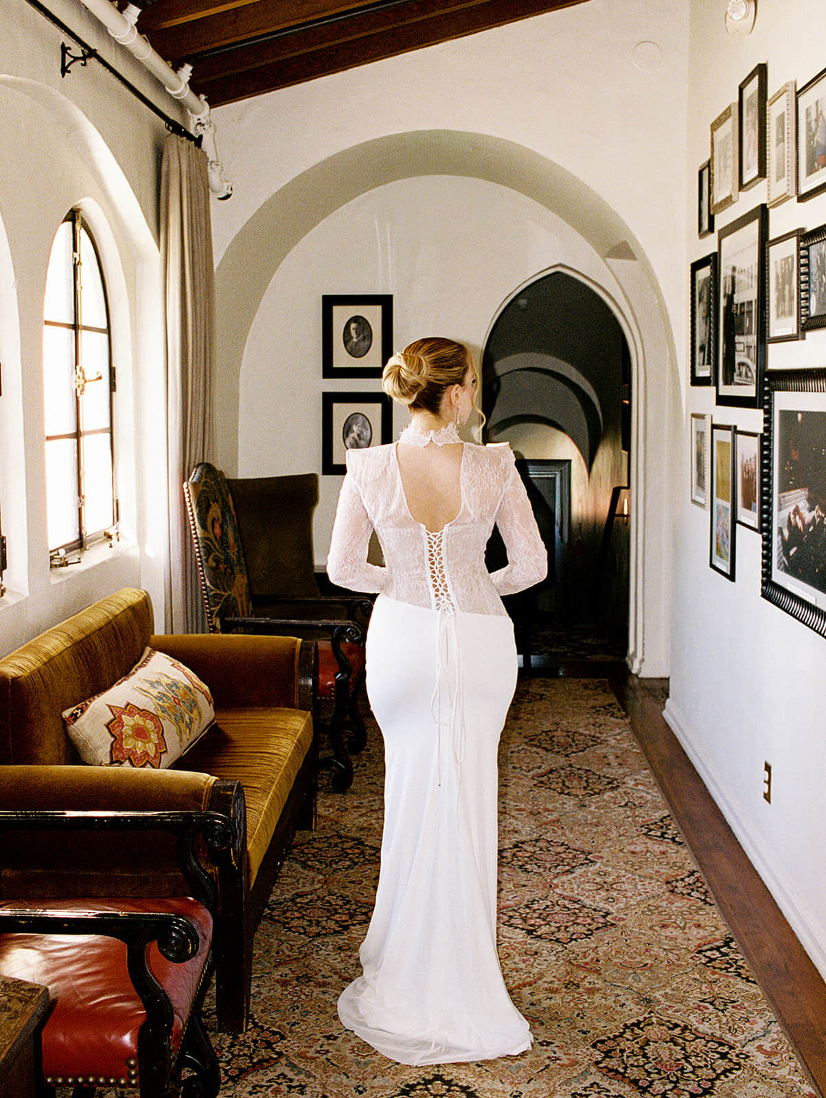 Bride standing in a softly lit, arched hallway, viewed from behind, wearing a fitted gown with a lace-up back and long sheer sleeves. Her hair is styled in an elegant low bun, and the space features a patterned rug, wood ceiling beams, framed artwork on the walls, and a cozy seating area by a window, creating a warm, timeless feel.
