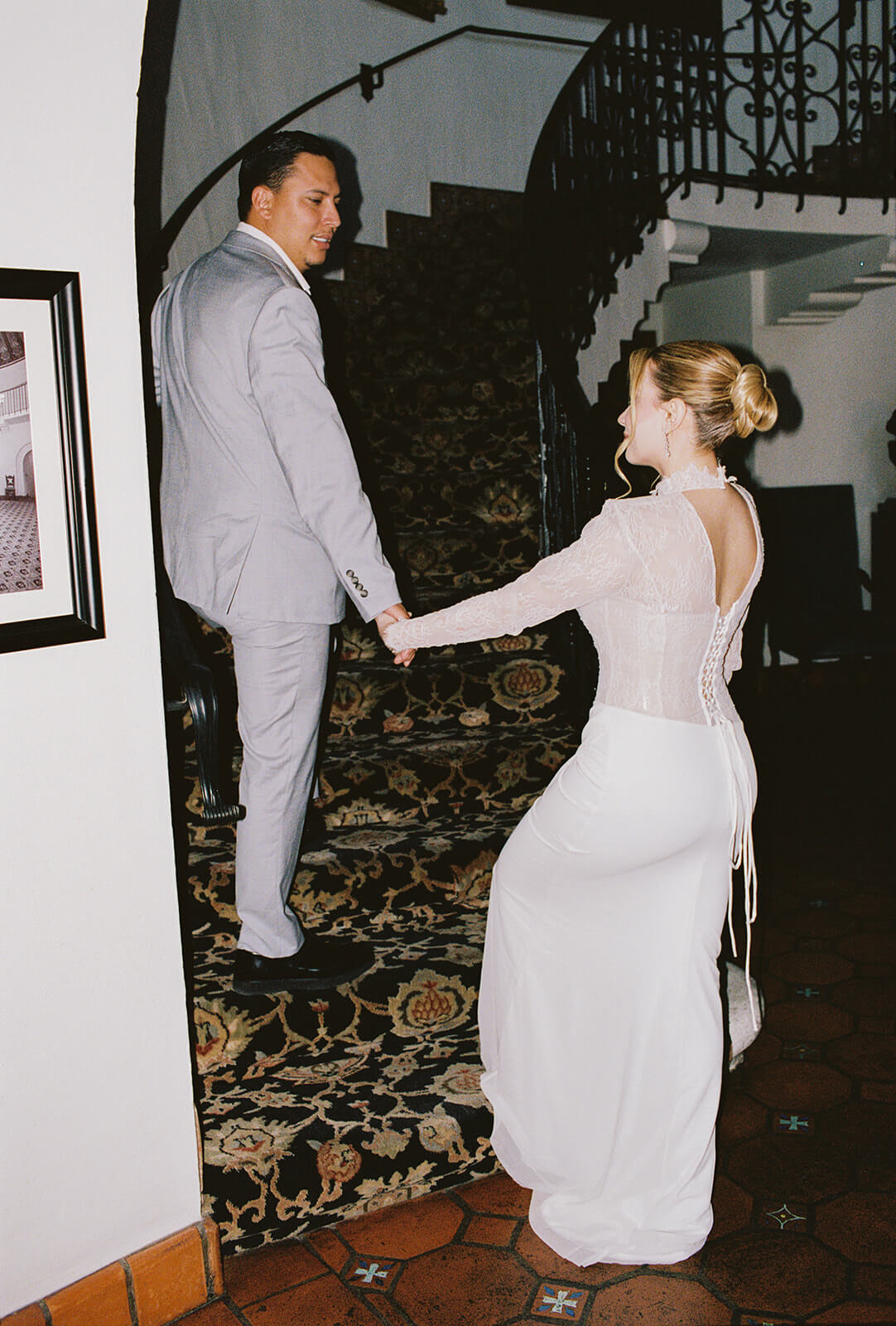 Bride and groom walking up a curved staircase inside an elegant micro wedding venue, holding hands as they move through a historic mansion with patterned carpet and wrought iron railing.