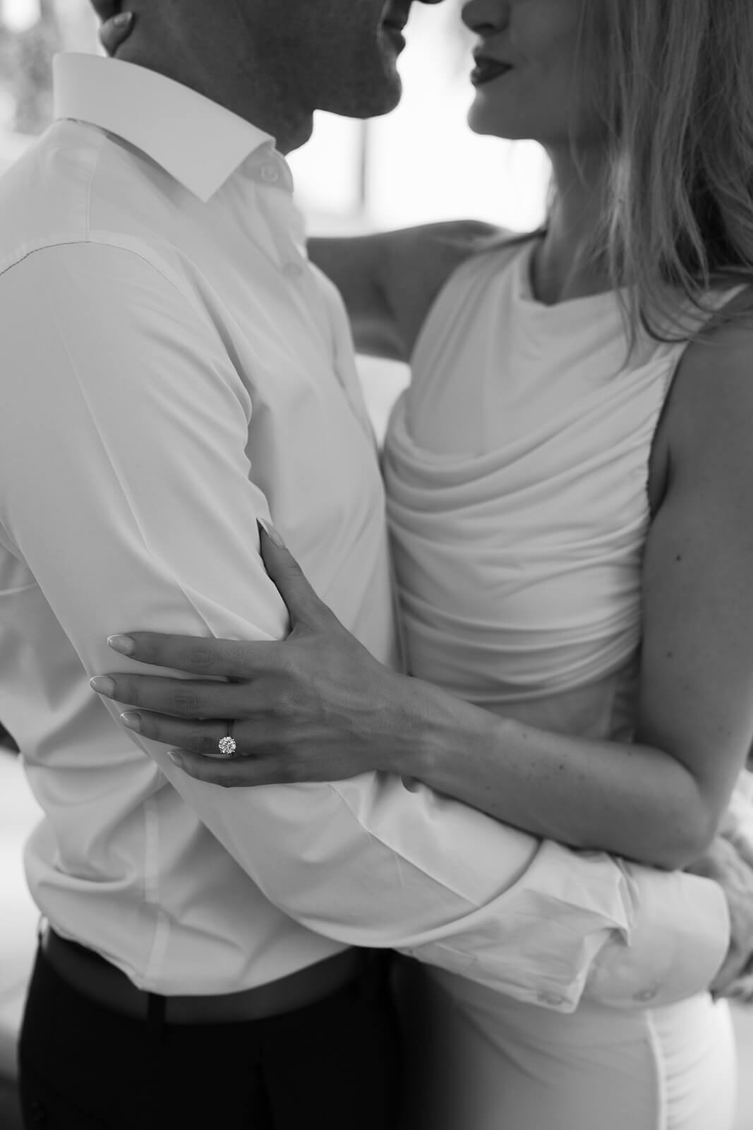 Close-up black and white detail of couple embracing, showing engagement ring and soft fabric of dress