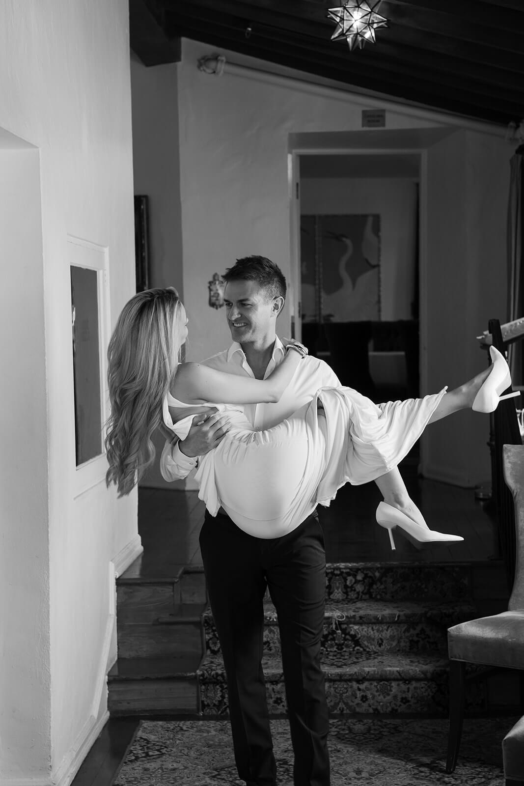Groom carrying bride in black and white photo inside Wrigley Mansion wedding venue in Phoenix Arizona with playful candid movement