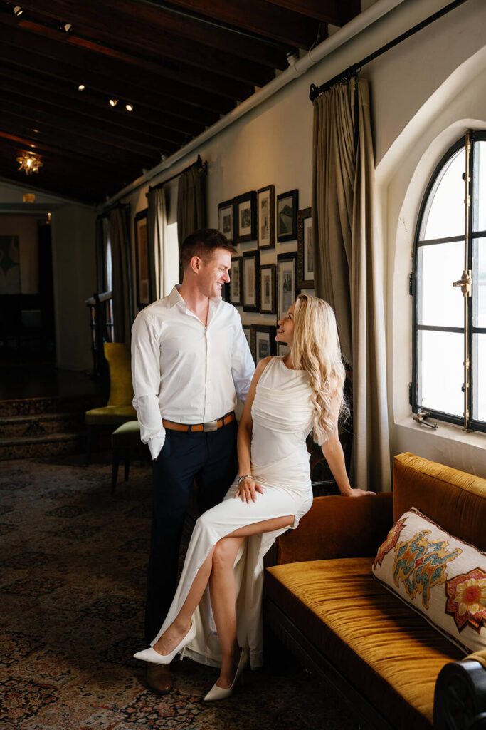 Couple sitting and standing near arched window at Wrigley Mansion wedding venue in Phoenix Arizona with warm sunlight and vintage interior details