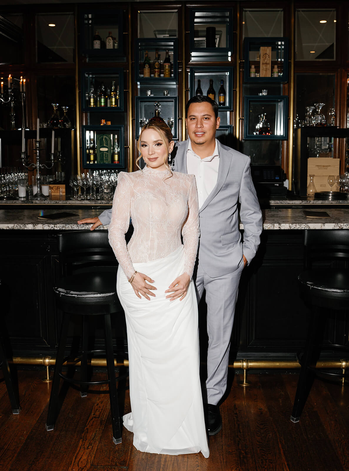 Bride and groom posing at a dark wood bar inside the Wrigley Mansion, an upscale Phoenix Arizona wedding venue, with glass shelves, liquor displays, and candlelight creating a moody, elegant backdrop.