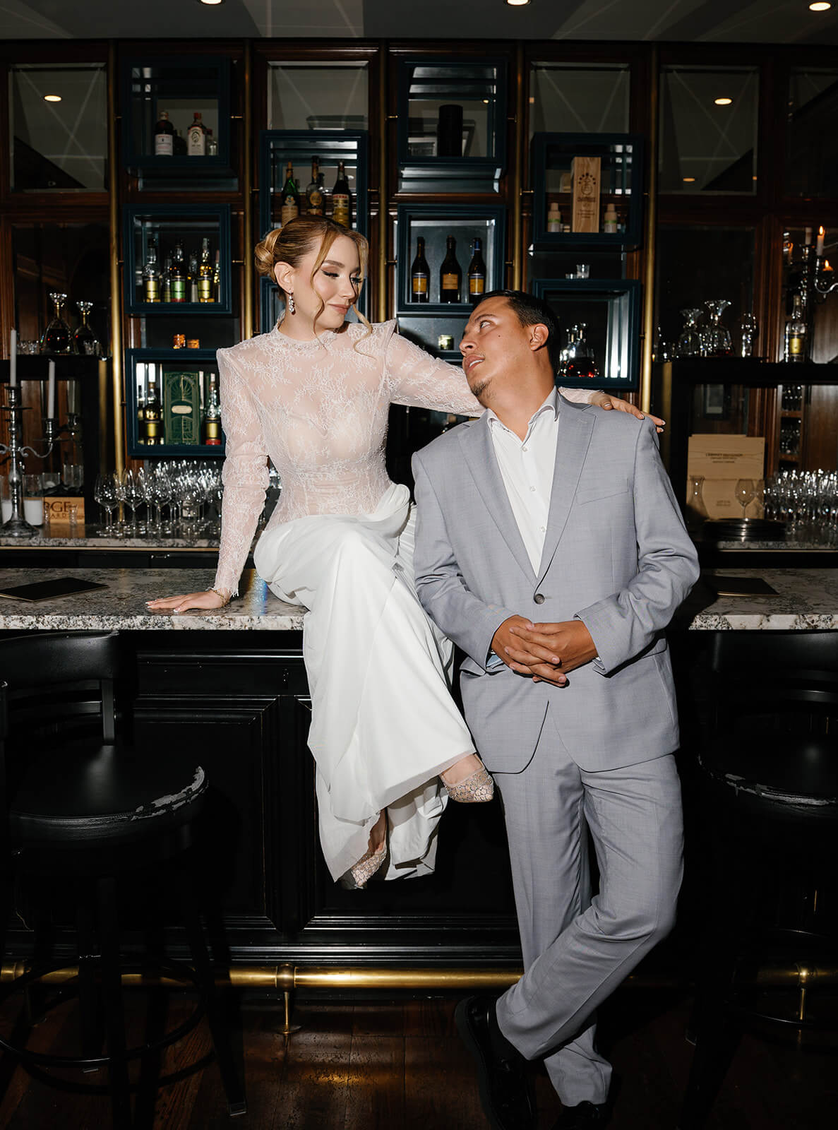 Bride sitting on the bar counter in a lace long-sleeve wedding dress while the groom leans beside her, sharing a playful moment at the Wrigley Mansion bar in Phoenix, Arizona.