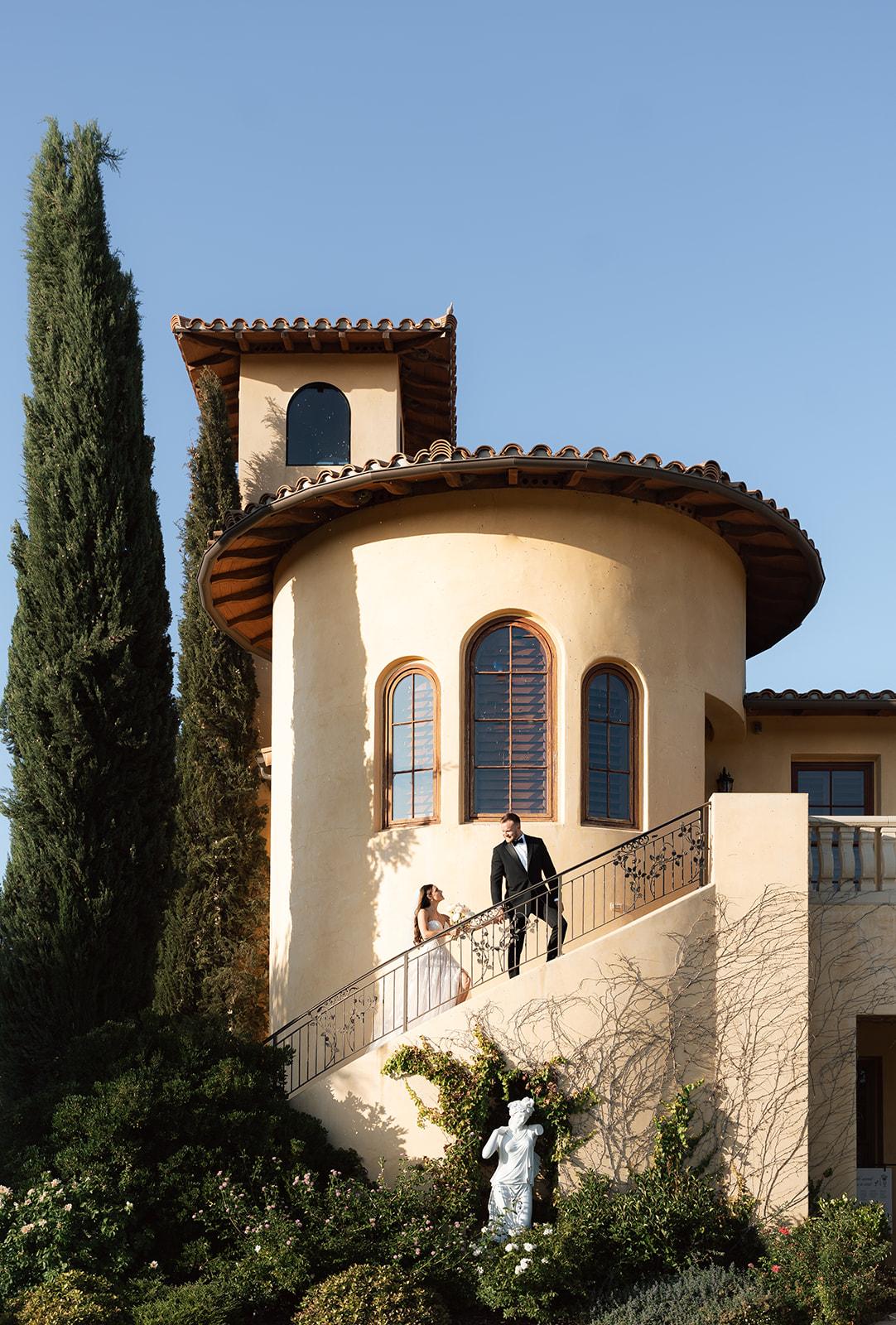 Couple walking down a curved staircase at Villa San Juliette, a Mediterranean-style wedding venue in SLO County with tall cypress trees and elegant architecture