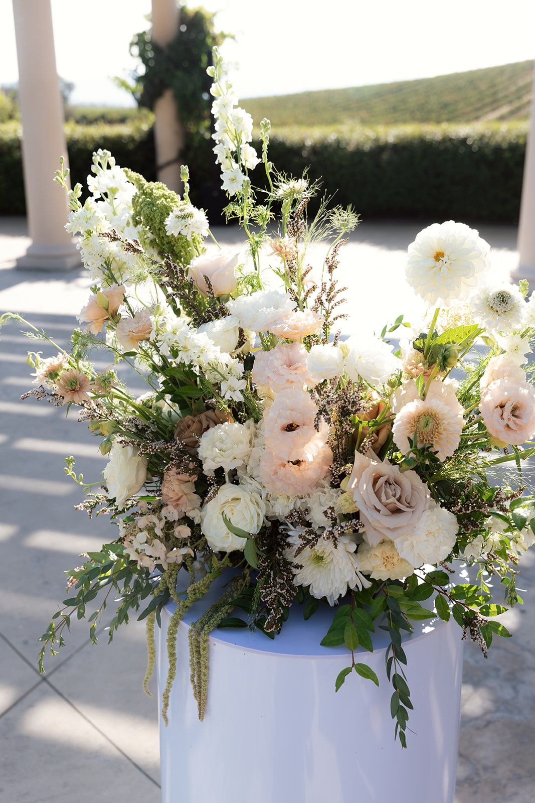 Lush floral ceremony arrangement of blush roses, white dahlias, and greenery on a white pedestal at an intimate wedding venue in California