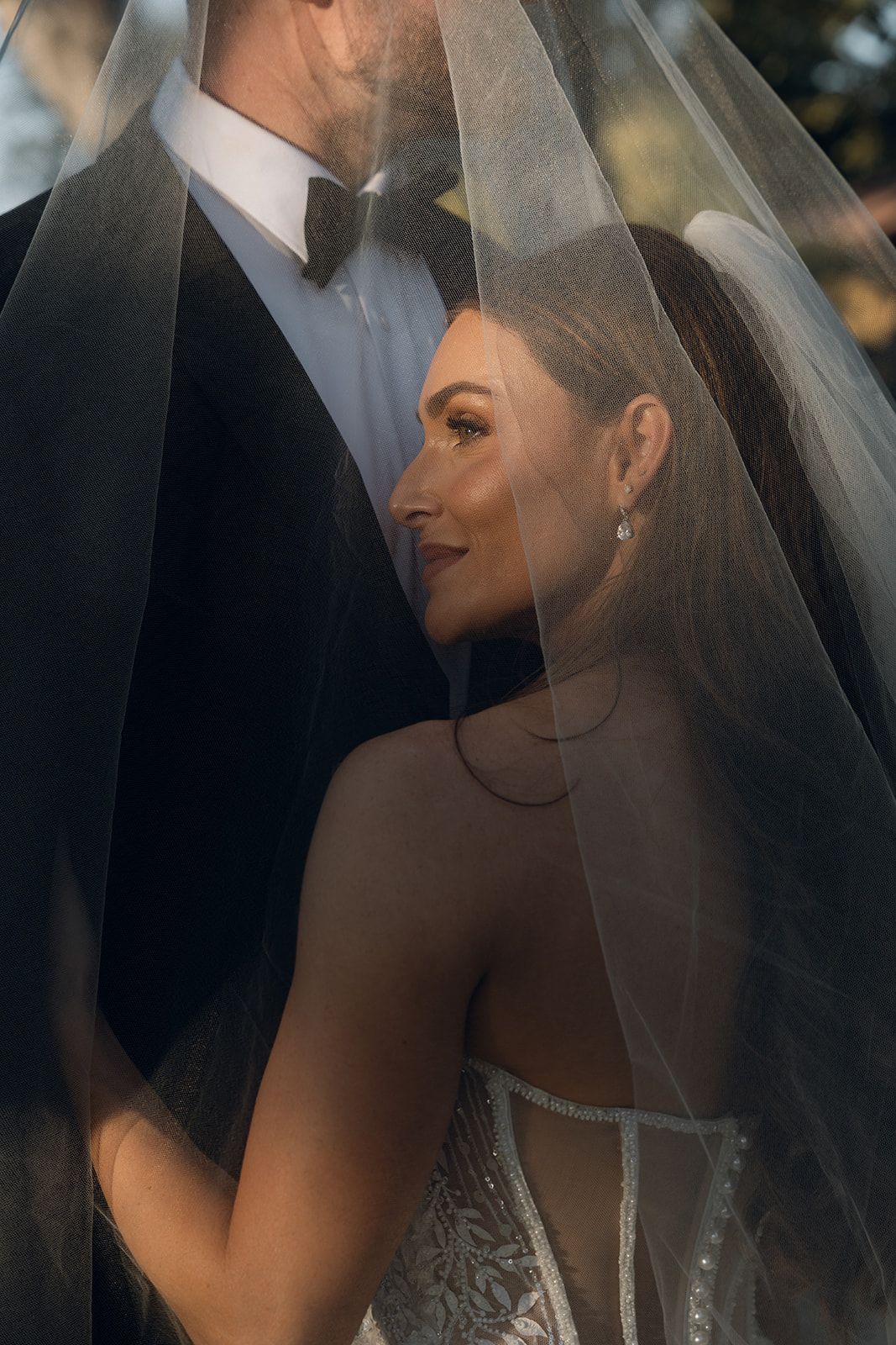 Romantic close-up portrait of a bride smiling beneath her flowing veil beside her tuxedo-clad groom at an intimate wedding venue in California