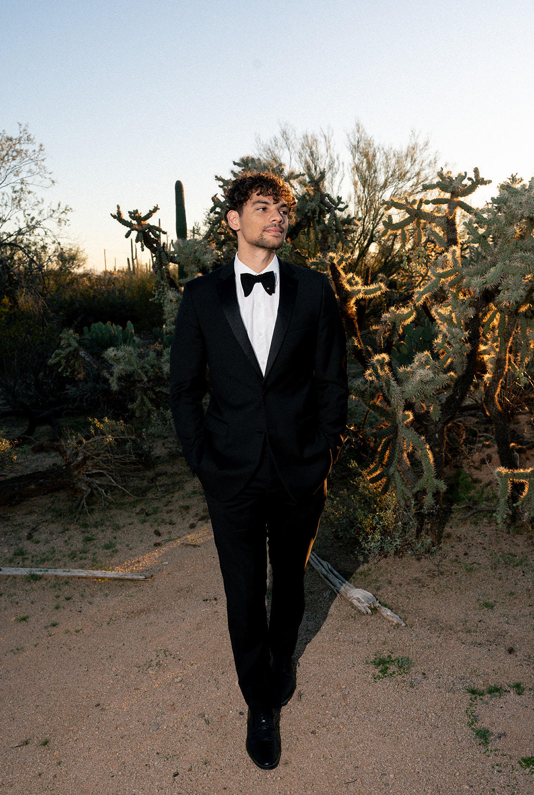 Groom stands alone in tuxedo among desert plants at golden hour