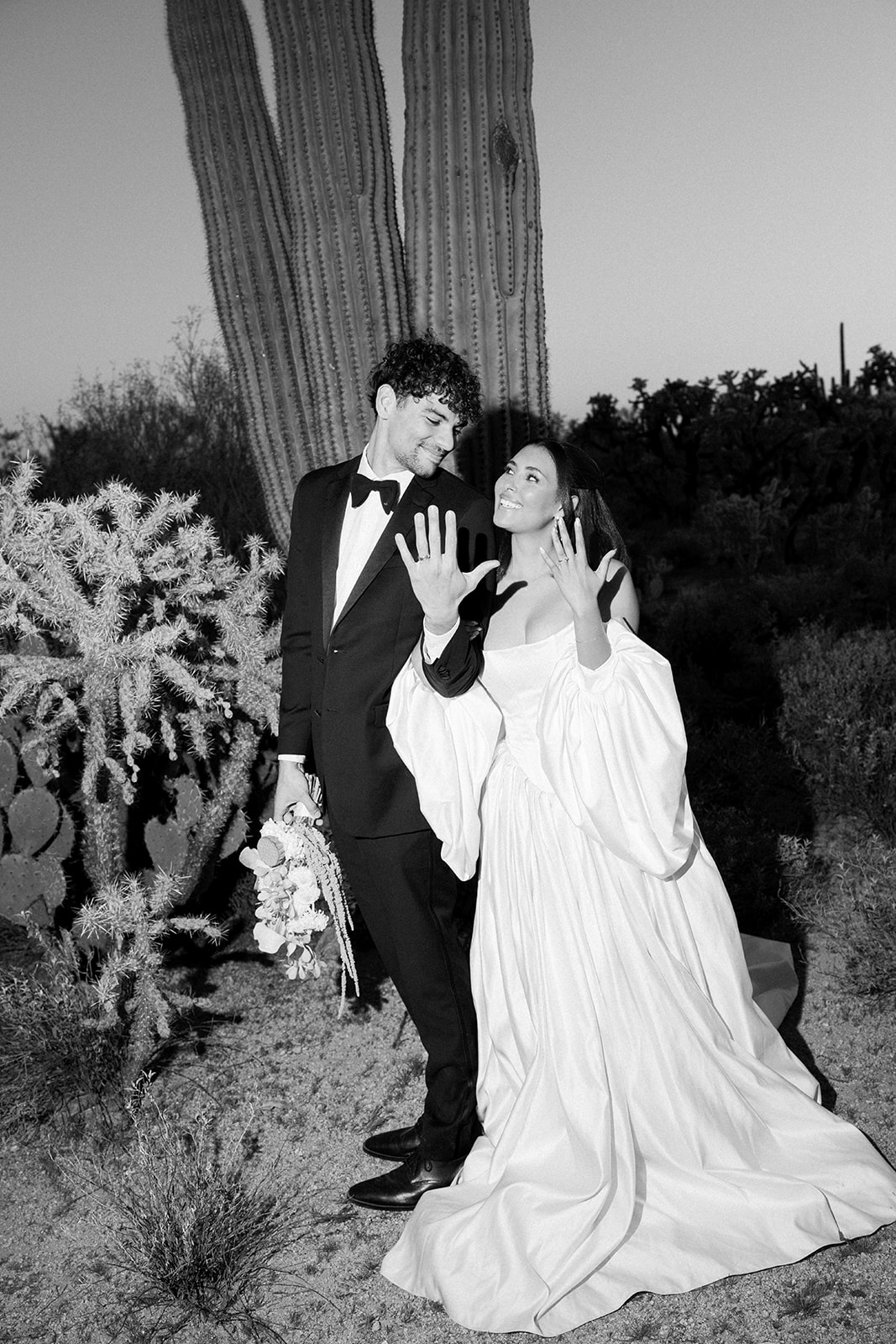 Bride shows ring beside groom among cacti in black and white