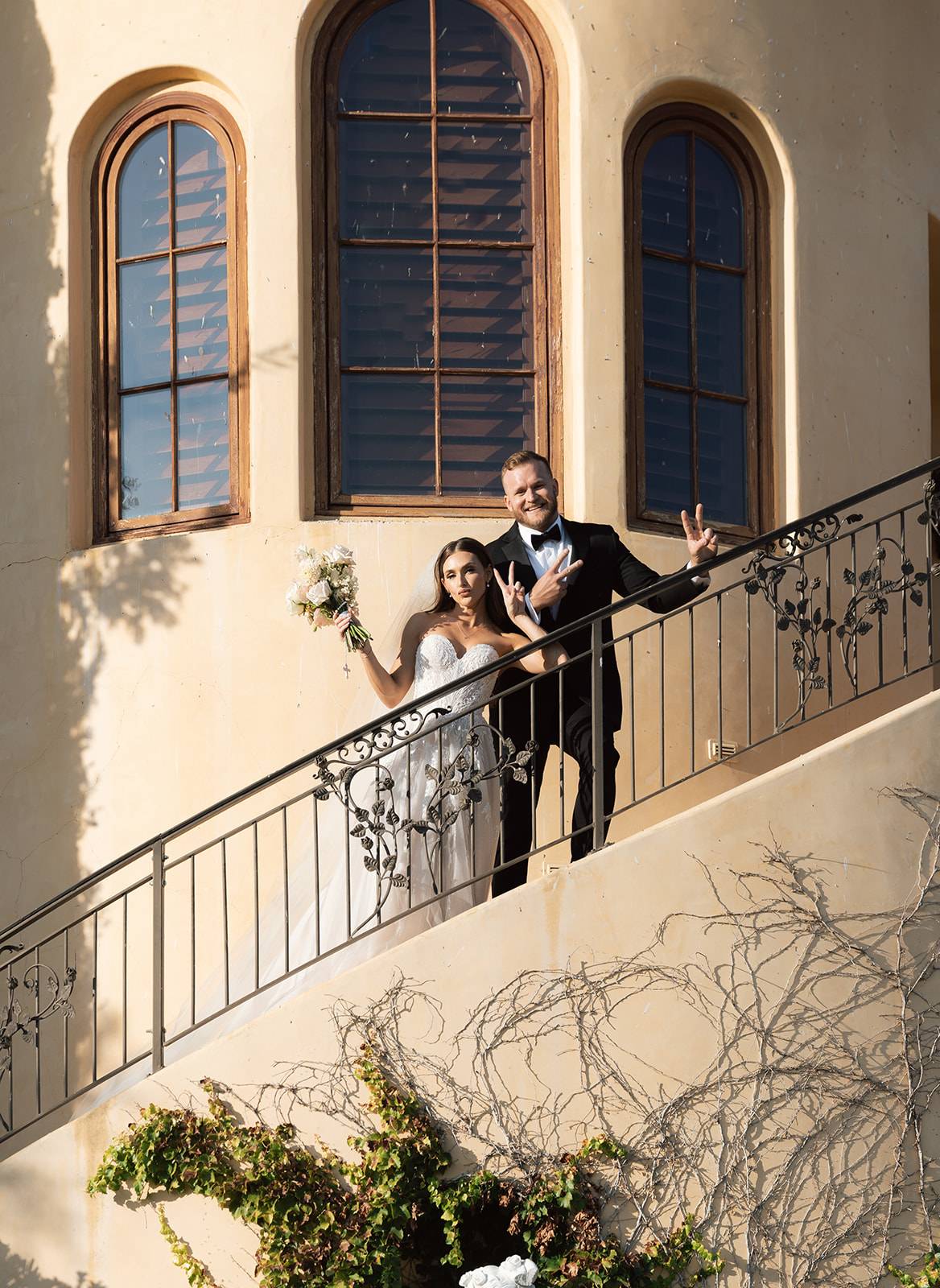 Bride and groom laughing and flashing peace signs from an ornate wrought iron balcony at a San Luis Obispo wedding venue