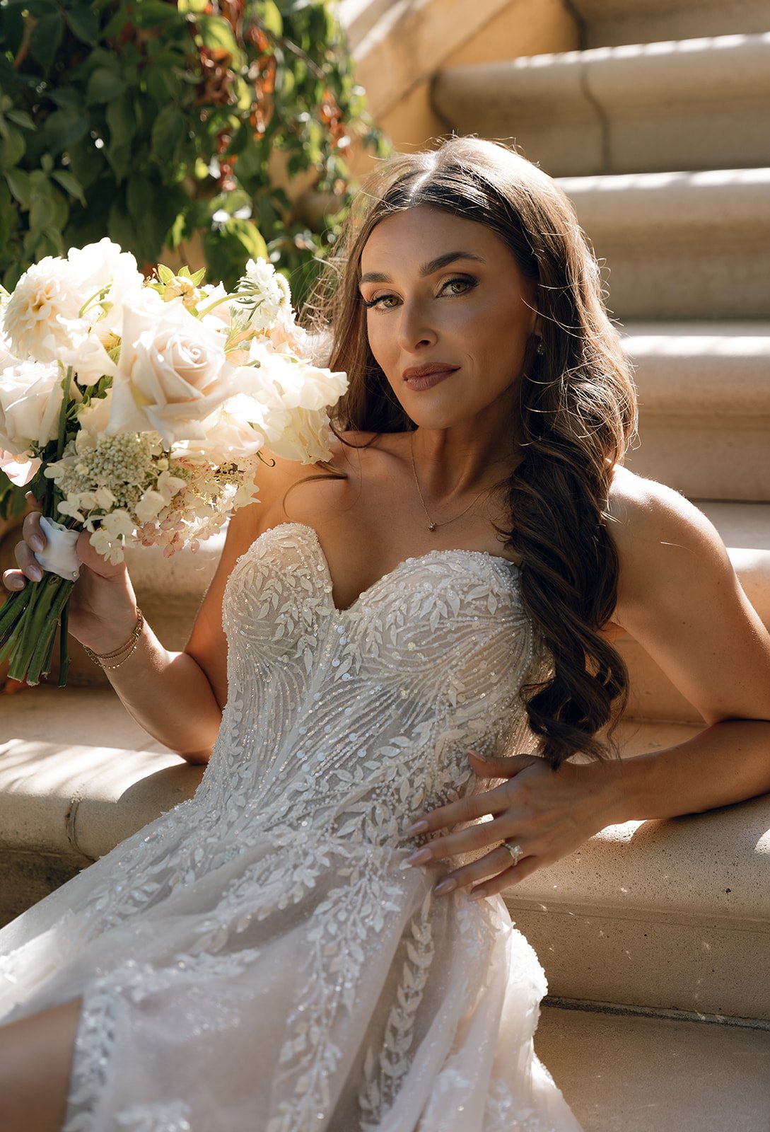 Bride seated on stone steps holding a soft blush and ivory bouquet, wearing a strapless beaded gown at a wedding venue in SLO county