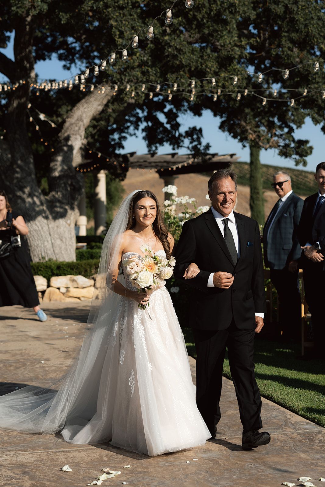 Smiling bride walking down the outdoor aisle with her father beneath string lights and oak trees at Villa San Juliette
