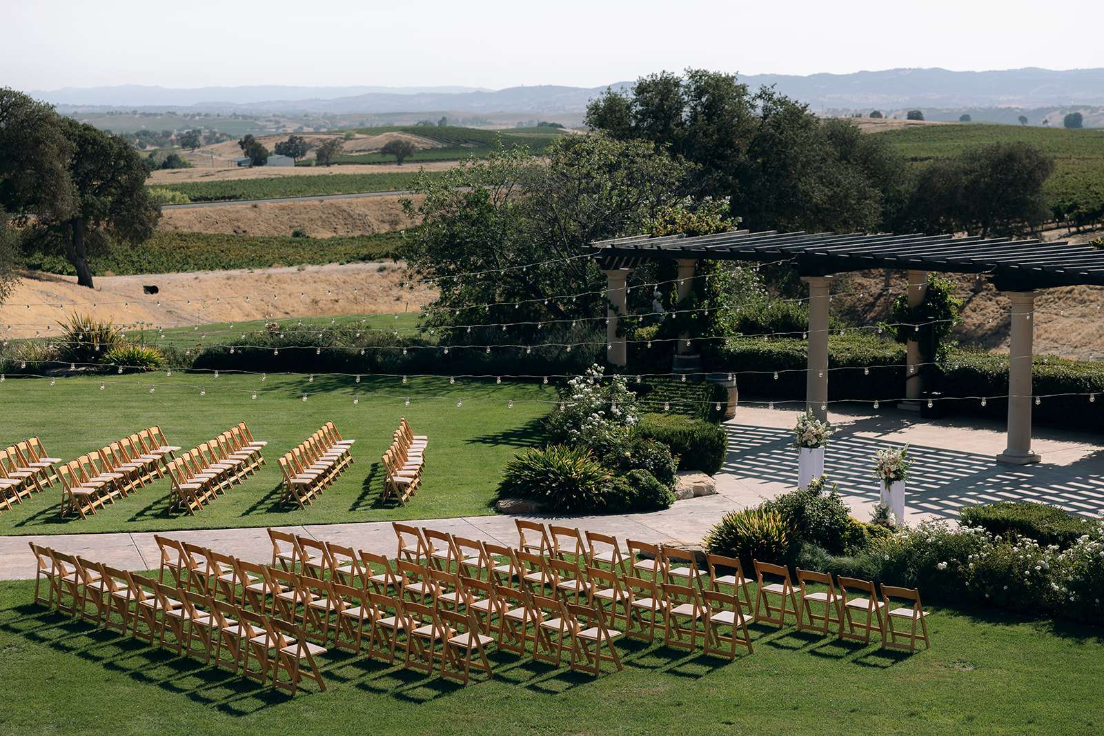 Aerial view of an outdoor wedding ceremony setup with rows of wooden chairs, string lights, and sweeping vineyard and mountain views at a San Luis Obispo wedding venue