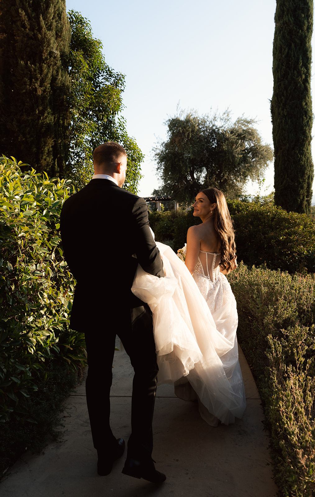 Groom carrying the bride's tulle train as she glances back with a smile while walking through the garden at a San Luis Obispo wedding venue