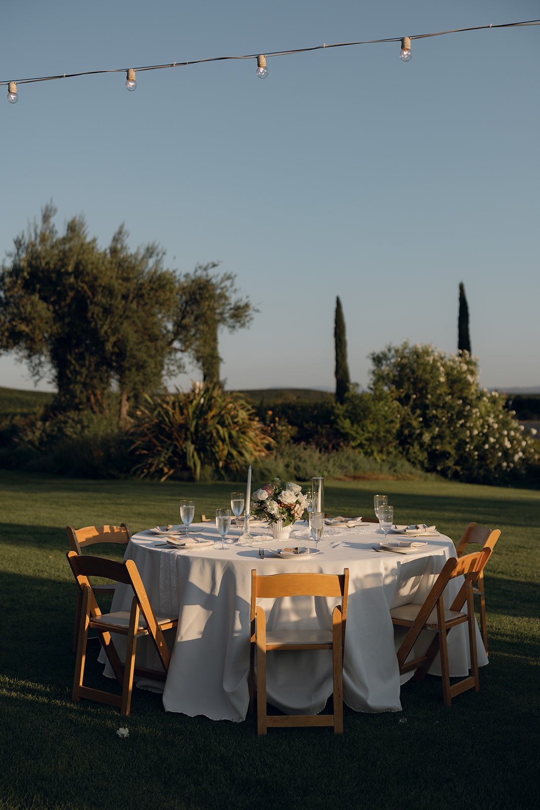 Elegantly set outdoor reception table with white linen, candles, and a floral centerpiece framed by cypress trees and open sky at Villa San Juliette