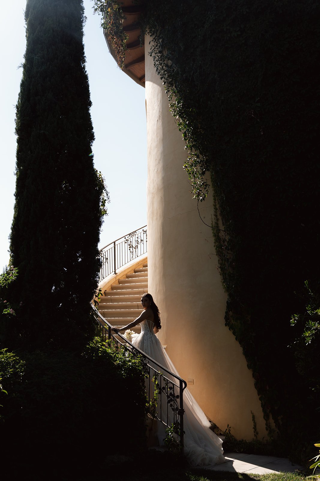 Dramatic bridal portrait of a bride descending an ivy-draped spiral staircase alongside a tall cypress tree at a San Luis Obispo wedding venue